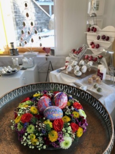 Bunt marmorierte Enteneier liegen in einem bunten Frühlingsblumenkranz. Im Hintergrund ist ein österlich, frühlingshaft dekorierter Verkaufsraum mit vielen Ostereiern zu sehen.